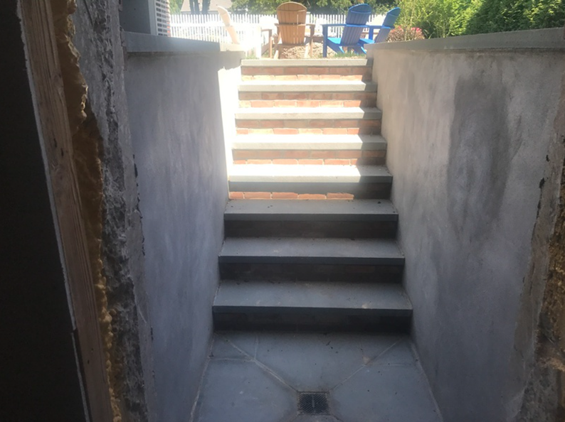 Stone steps leading up from a concrete basement area to an outdoor patio with blue chairs.
