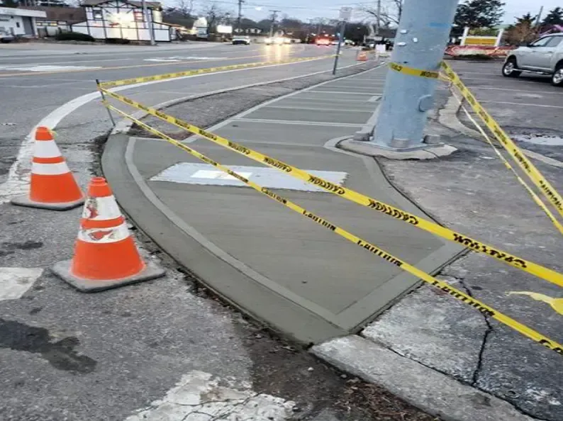 Fresh concrete curb with caution tape and cones near a road and a tree.