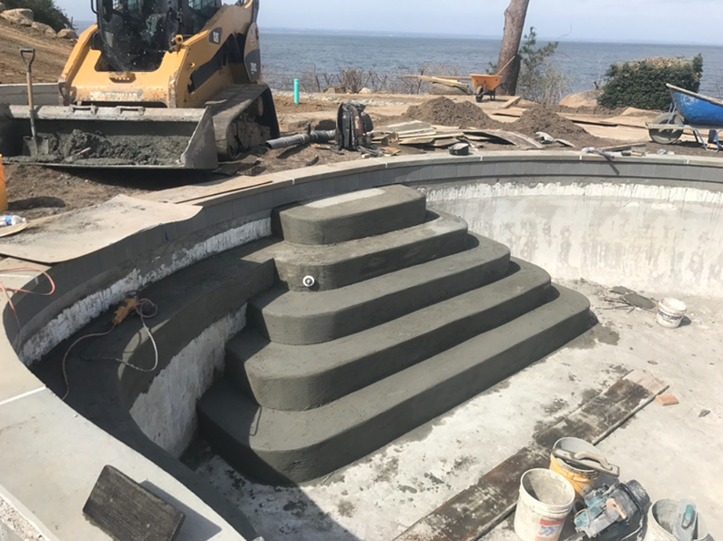 Construction of a concrete pool with steps; a skid steer in background; setting is outdoors.