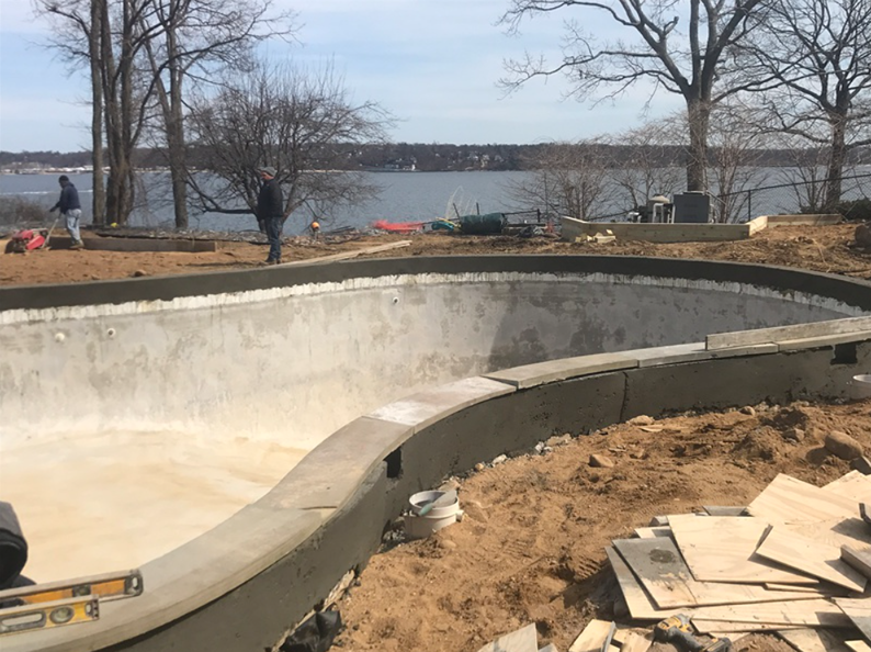 Construction of an empty concrete pool bowl near a body of water; workers nearby.