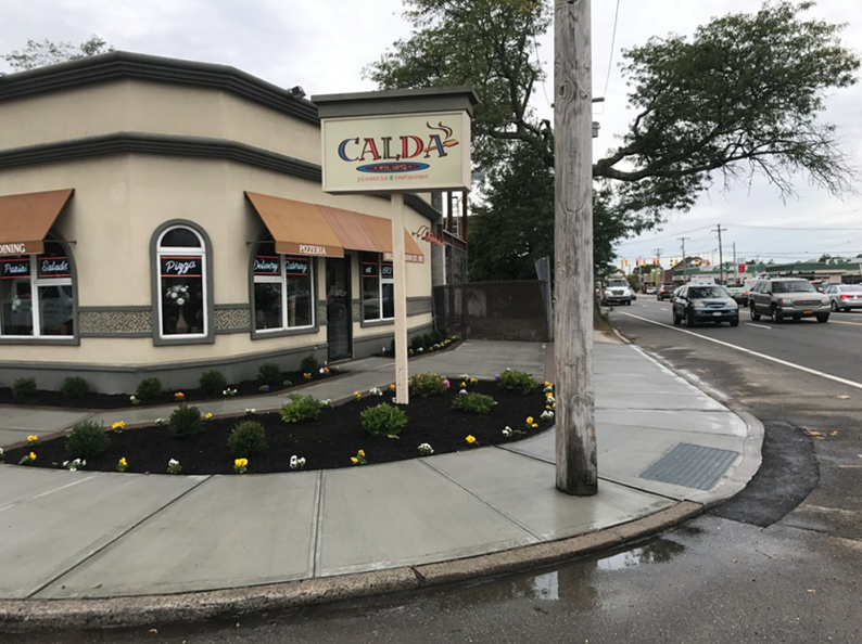 Corner building, CALDA Restaurant sign, brown awnings, flowers, sidewalk, cars on street.