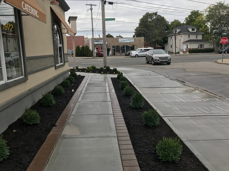 Sidewalk with bushes, leading to a street corner with cars and buildings under a cloudy sky.