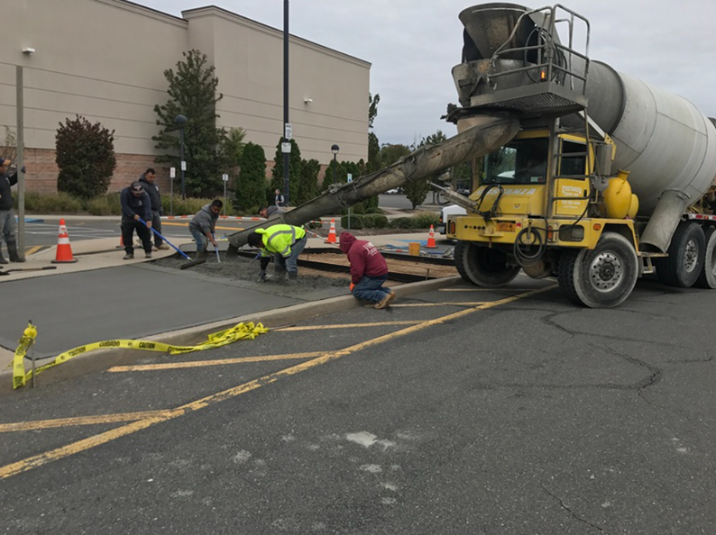 Workers pouring concrete from a yellow truck to pave a road, with yellow caution tape nearby.