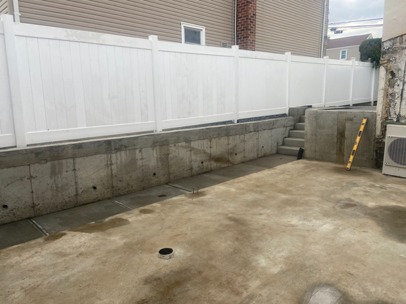 Concrete patio with a retaining wall, steps, and white fence. A level leans against a wall.