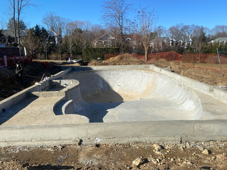 Concrete skate bowl under construction in an outdoor setting, with surrounding dirt and residential homes.