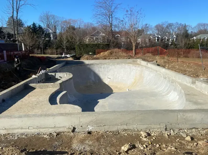 Construction site with a concrete skate bowl; surrounding dirt and fencing; homes in background.