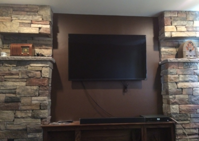TV mounted on brown wall between stone columns; soundbar and components below.