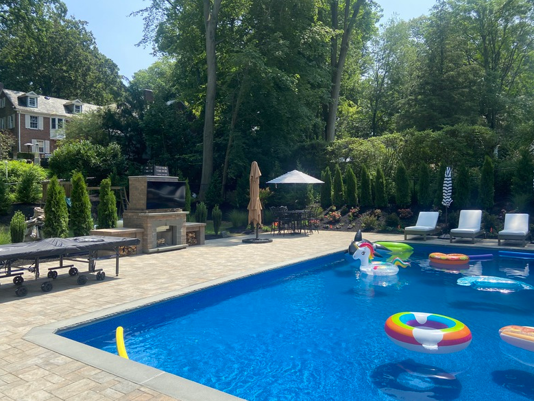 Backyard pool with inflatables. Lounge chairs, umbrella, and a TV stand are on the paved deck.