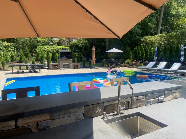 Backyard pool with umbrella, bar, lounge chairs, and grill. Bright blue water.