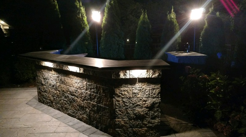 Outdoor bar with stone facade illuminated at night. Overhead lights shine onto the counter and foliage.