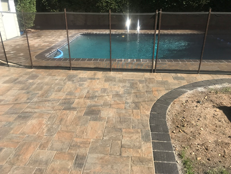 Paver patio next to a pool with safety fence; brown and black bricks, blue water, sunny outdoor setting.