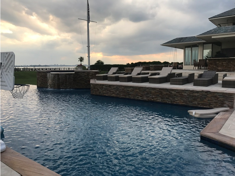 A pool with lounge chairs, overlooking a body of water, next to a house, under a cloudy sky.