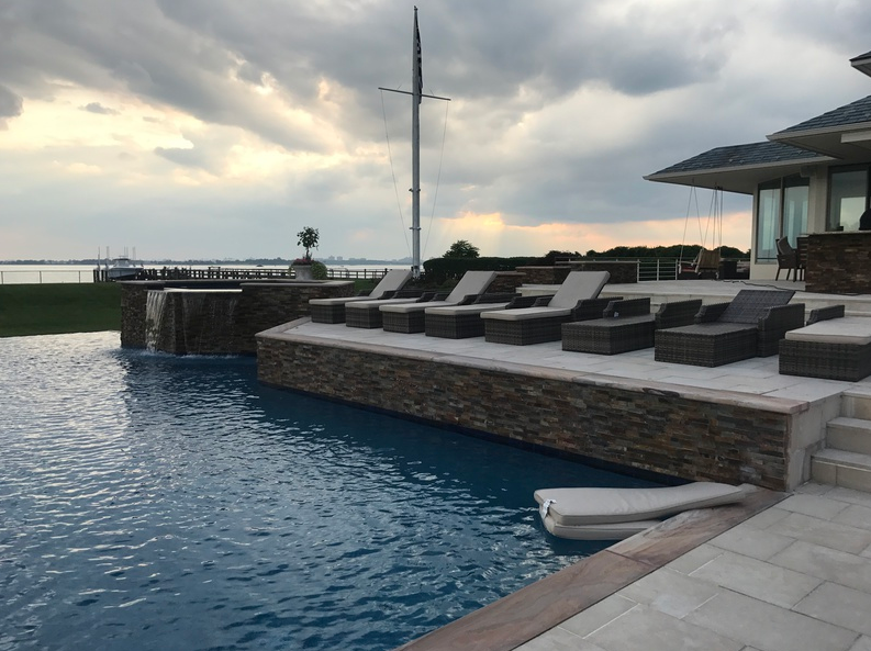 Infinity pool next to a house with lounge chairs overlooking a body of water under a cloudy sky.