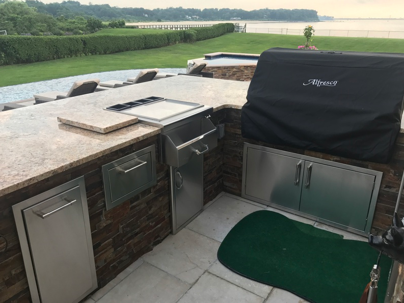 Outdoor kitchen with grill, cabinets, and a green mat, near a body of water.