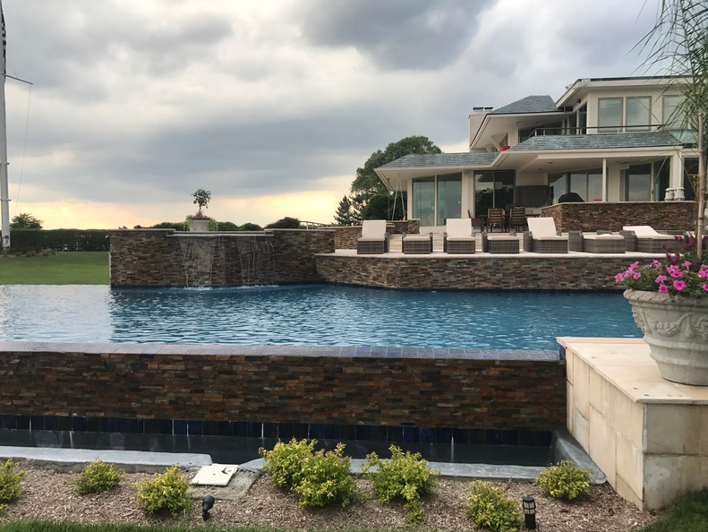 Luxury home with infinity pool, waterfall feature, and outdoor seating against a cloudy sky.