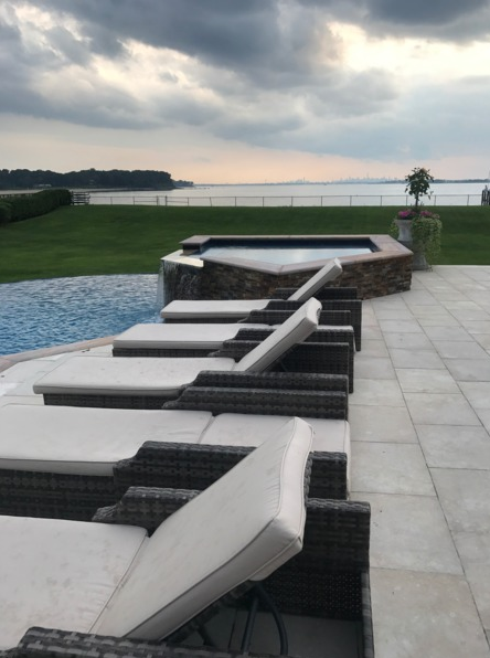 Poolside scene: loungers, infinity pool, spa, overlooking water under cloudy sky.