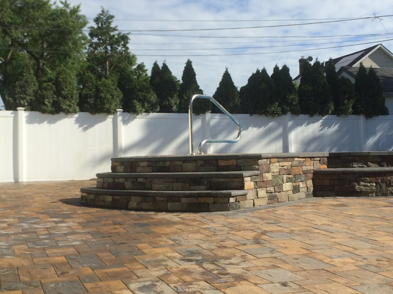 Stone steps with handrail lead to a raised stone structure, set on a brick patio, near a white fence.