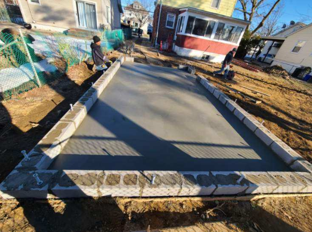 A freshly poured concrete slab surrounded by cinder blocks in a residential yard. People work nearby on a sunny day.