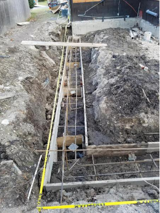 Foundation construction site: trenches with rebar framework, wooden supports, and piles of dirt.
