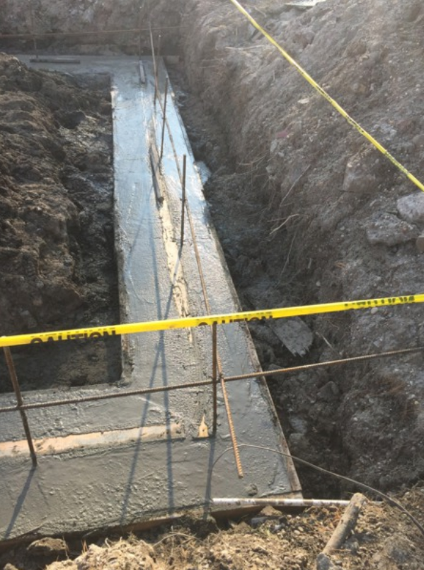 Newly poured concrete foundation with rebar in an excavated trench, marked with yellow caution tape.