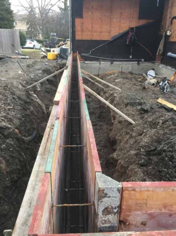 Wooden forms and rebar in a trench, prepared for a concrete foundation pour beside a building.
