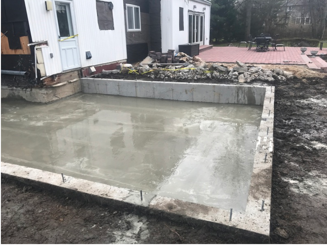 A concrete patio under construction next to a white house with a red brick patio.