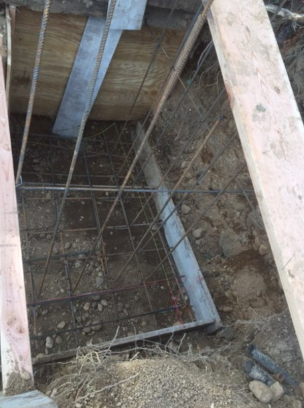 Construction site: steel reinforcement cage for concrete foundation within a wooden frame, set in dirt.