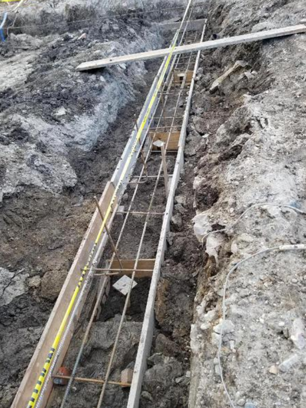 A narrow trench with metal rebar framework inside, ready for concrete, surrounded by dirt and wooden planks.