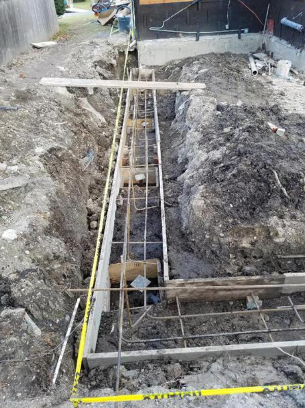Construction site trench with rebar framework, awaiting concrete. Dirt, yellow caution tape, and wood visible.