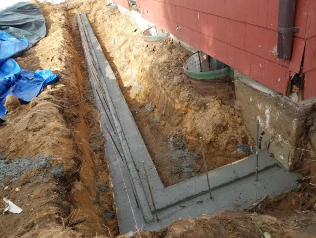 Foundation repair: trench dug along red building's side, with new concrete and rebar forming a base.
