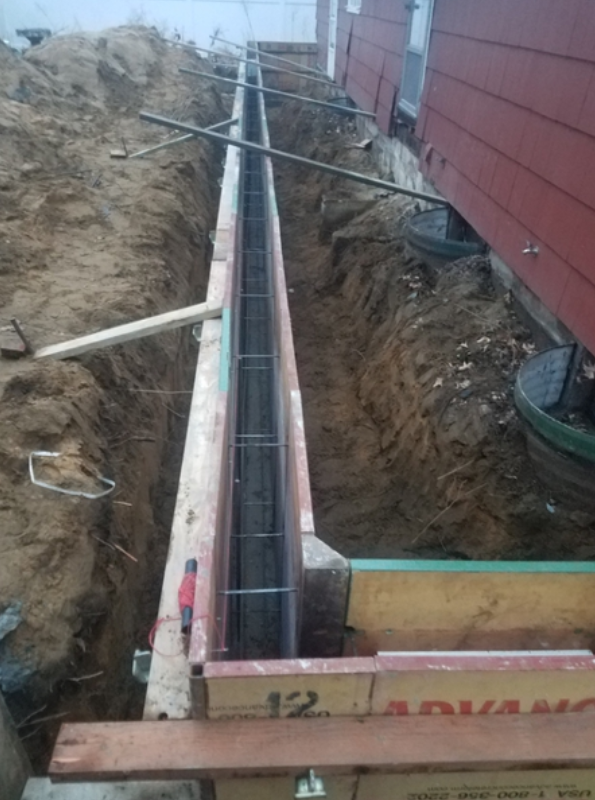 Construction site: trench with wooden forms, steel rebar, and soil near a building.