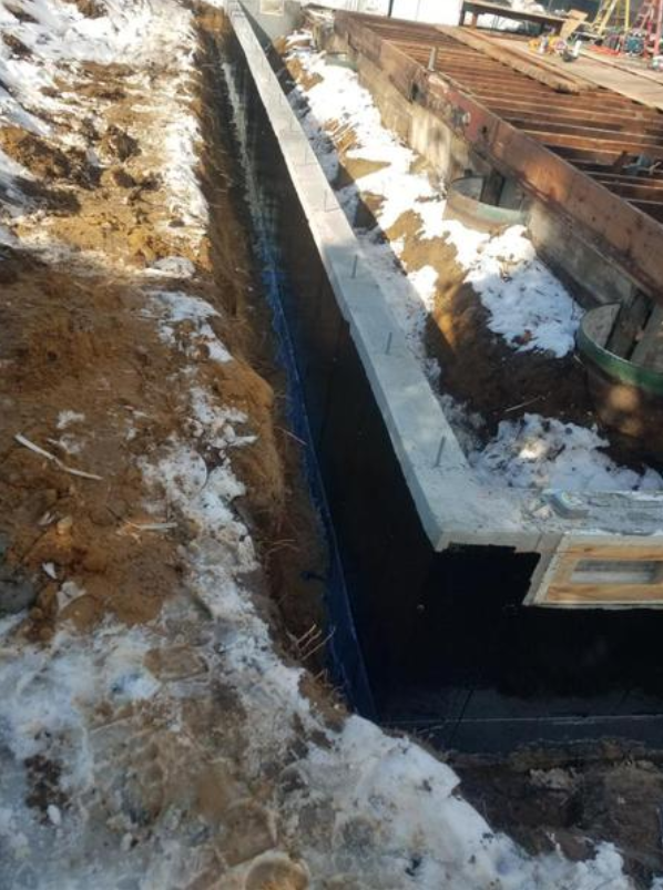 Trench dug next to a wooden structure with snow, concrete, and exposed earth.