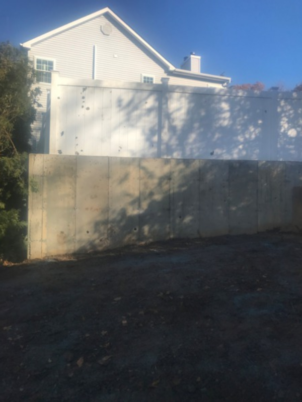 A concrete retaining wall in front of a white house under a blue sky, with tree shadows.
