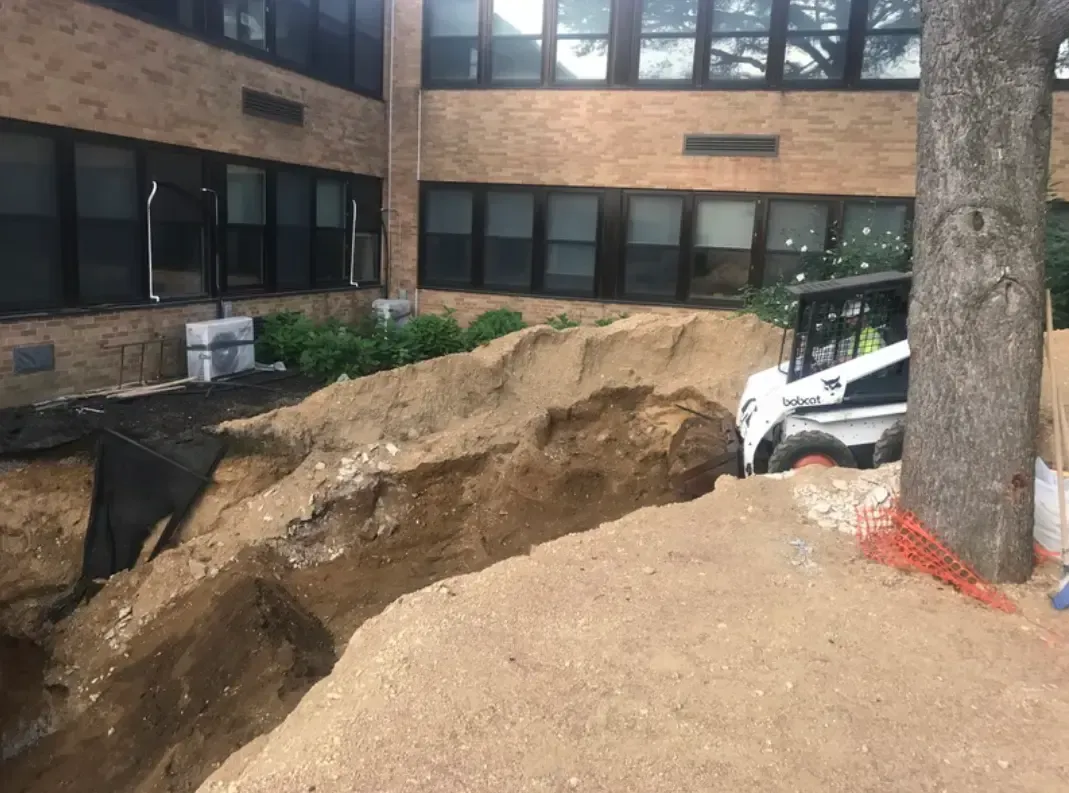 Construction site near brick building: trench, dirt pile, small bulldozer, and a tree.