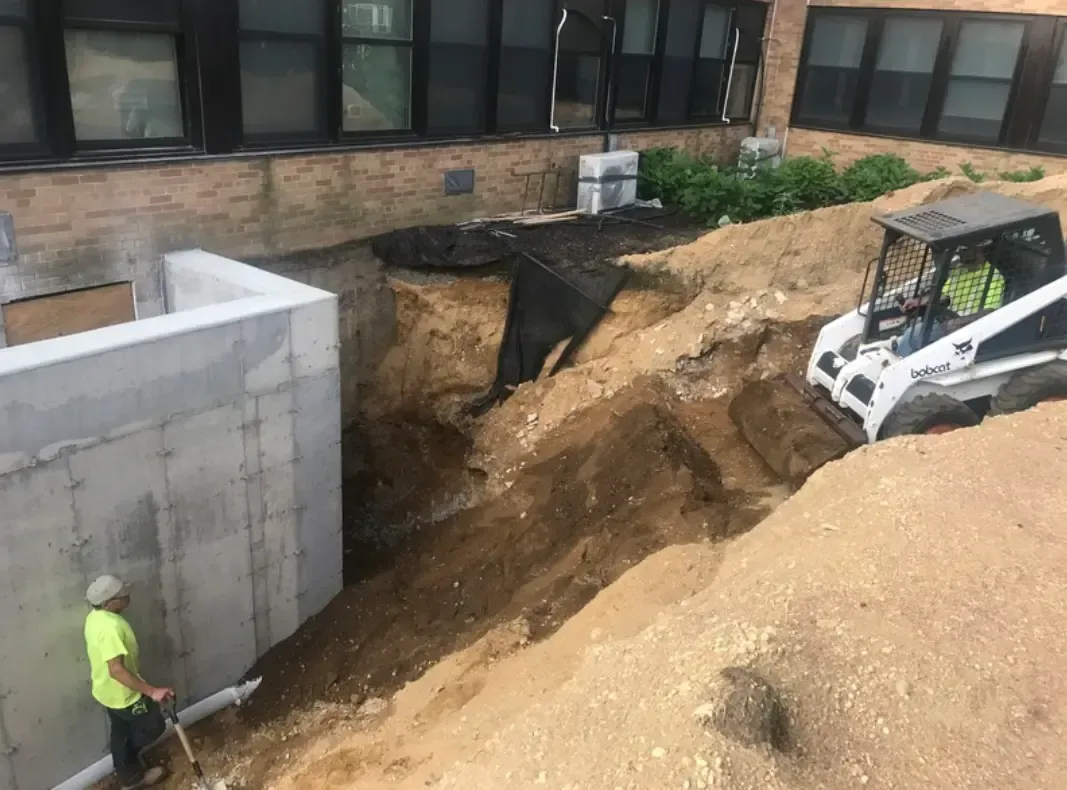 Construction site: Worker near concrete wall; bobcat excavates soil near building.