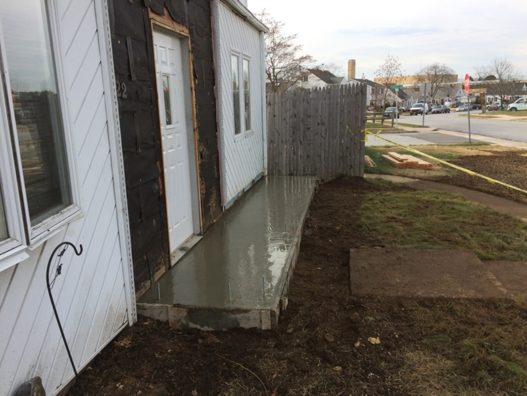 Freshly poured concrete slab porch outside a white house, next to a grass yard and street.