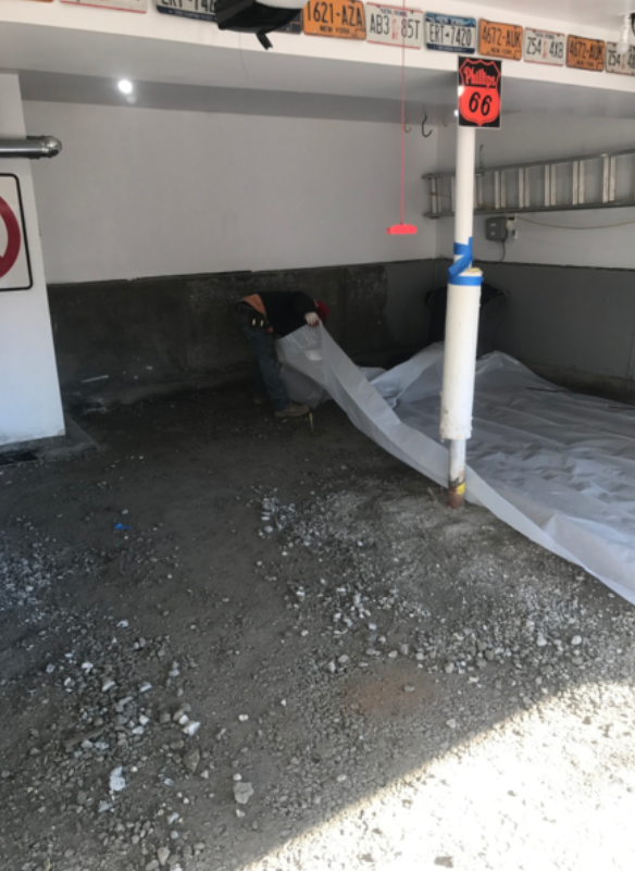 Person in a garage, working on the floor. Gravel and plastic sheeting are present. A white pipe is also visible.