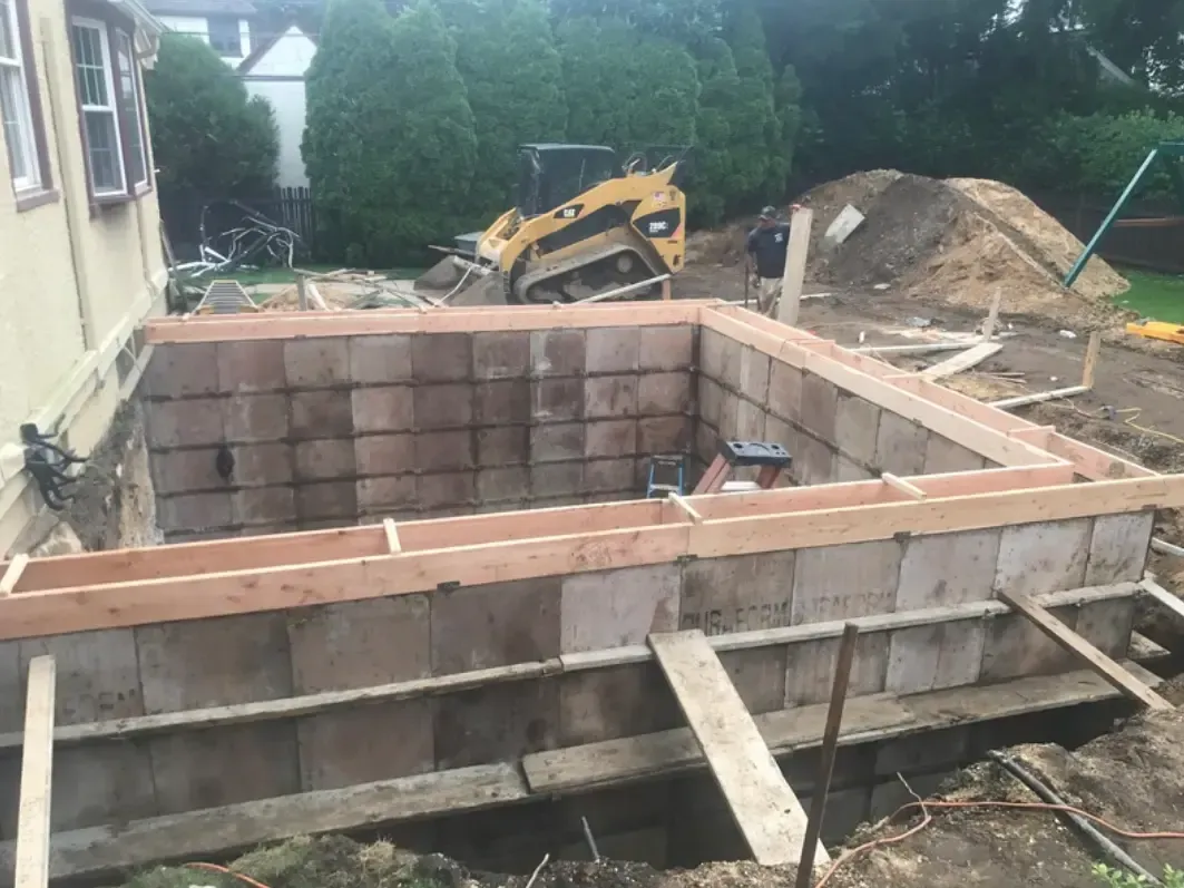 Construction site with a rectangular concrete block structure, wooden framing, and a small excavator.