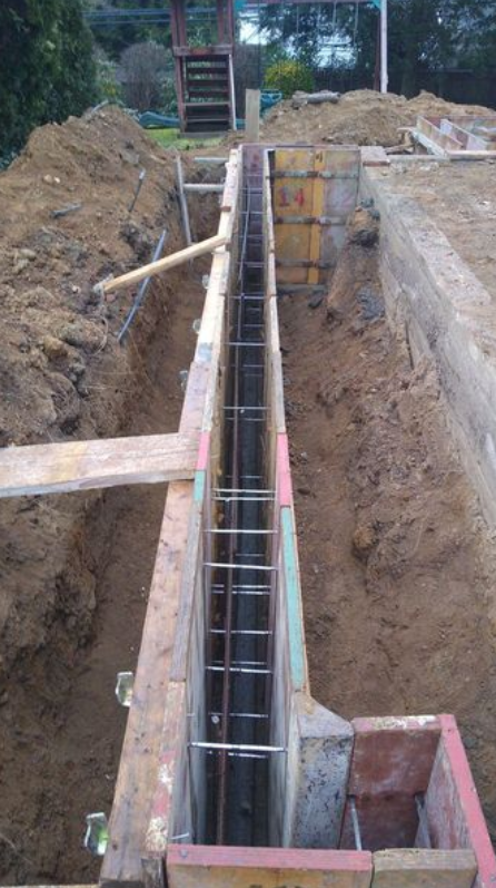 A rectangular trench dug into soil with concrete foundation forms, rebar framework, and wooden supports.