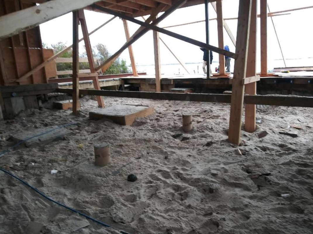 Construction site under a wooden structure, with sand on the ground and exposed beams.