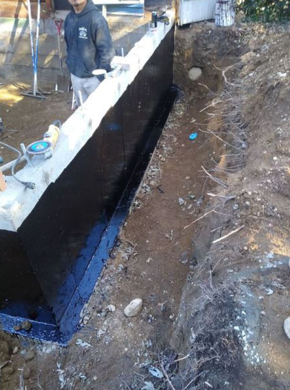 Construction worker near a concrete foundation wall, black waterproof membrane applied, in a dirt trench.