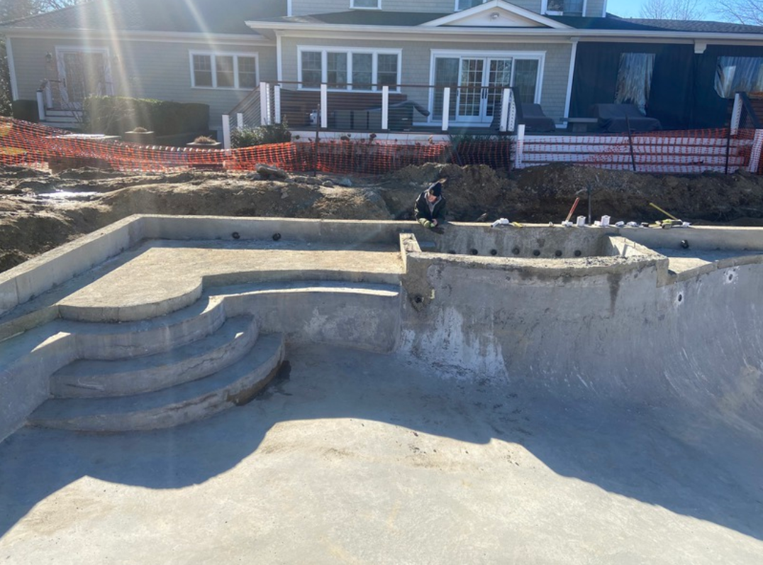 Concrete pool construction site with steps and a raised spa, in front of a house.