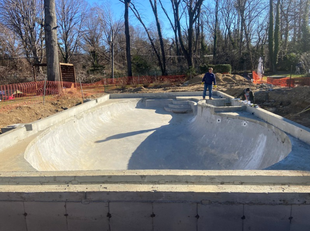 A concrete skate bowl under construction. A person stands inside, surrounded by trees.