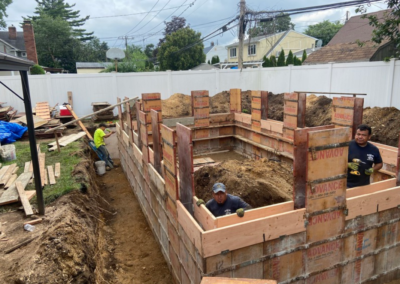 Construction workers building a brick foundation outdoors, with a trench, tools, and lumber visible.