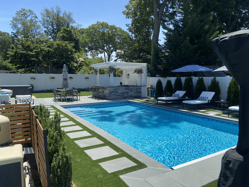 A backyard pool with lounge chairs, a pergola, and an outdoor kitchen, surrounded by green grass and trees.