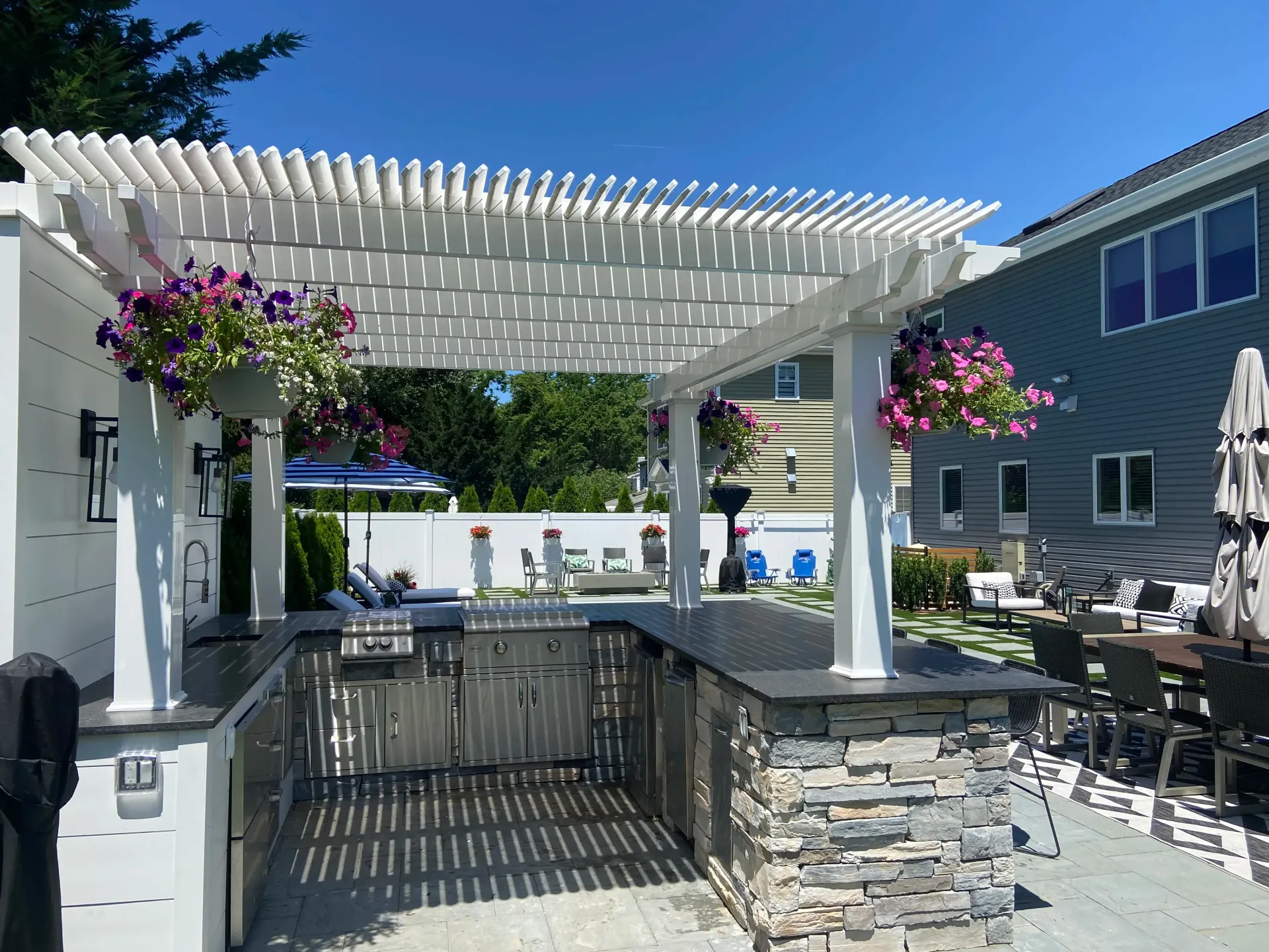 Outdoor kitchen with white pergola, stone accents, grill, and hanging flower baskets.