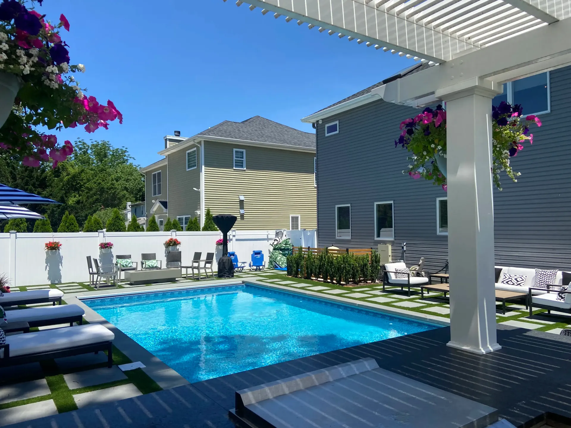 Pool with lounge chairs, pergola, and hanging flower baskets. Houses and a sunny sky in the background.