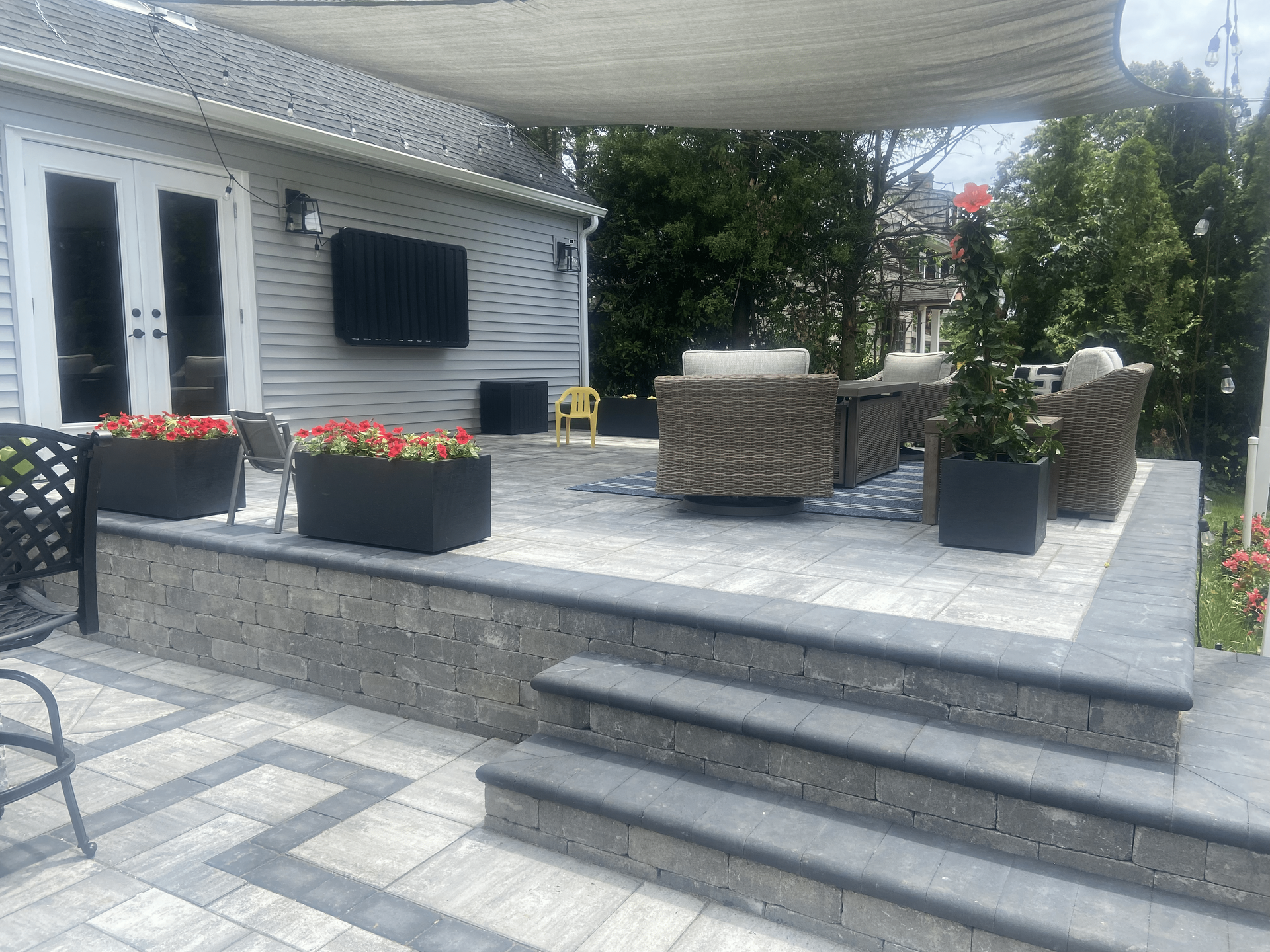 Patio with pavers, steps, and seating. Gray and brown tones, flowers, and a house in the background.