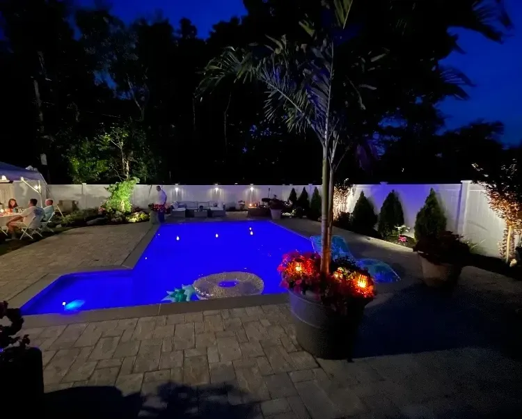 Nighttime view of a pool, illuminated blue, surrounded by patio, garden, and lit white fence.