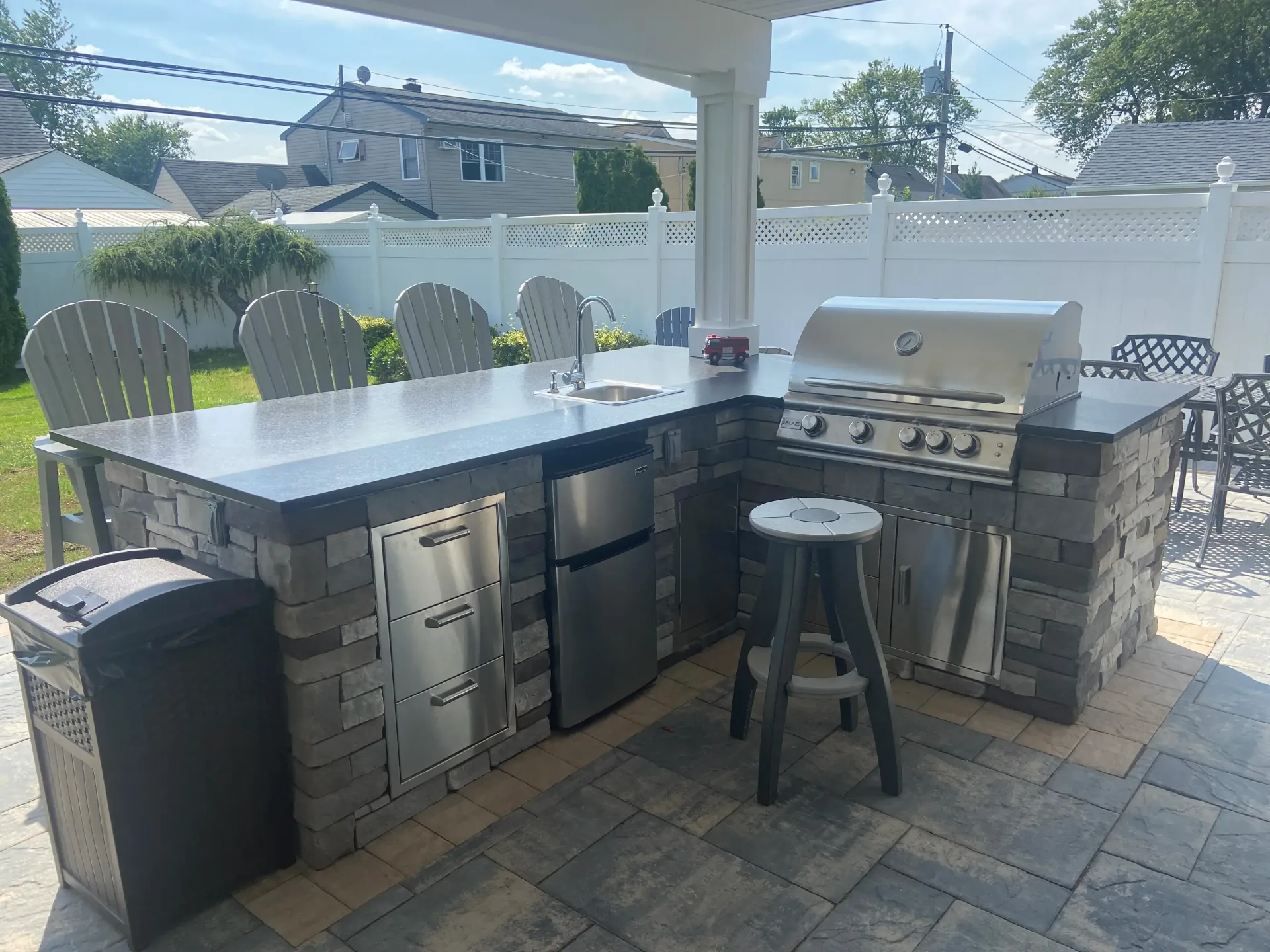 Outdoor kitchen with a grill, sink, refrigerator, and seating, under a white pergola.